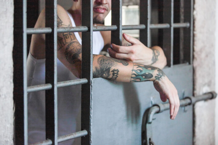 Osoba w białym podkoszulku opiera ramiona o żelazne pręty okna, ukazując tatuaże o różnych wzorach na przedramionach