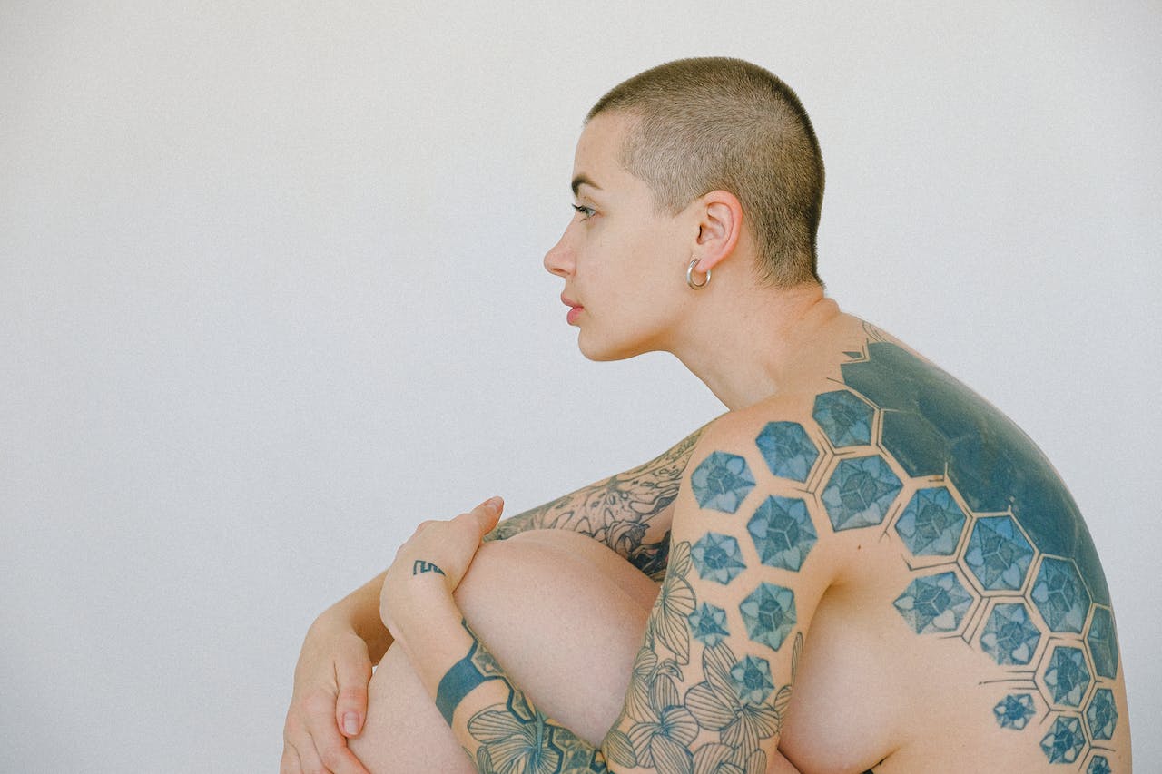 Kobieta z krótkimi włosami siedzi skulona, prezentując tatuaże pokrywające jej ramię i plecy, w tym geometryczne wzory i kwiaty