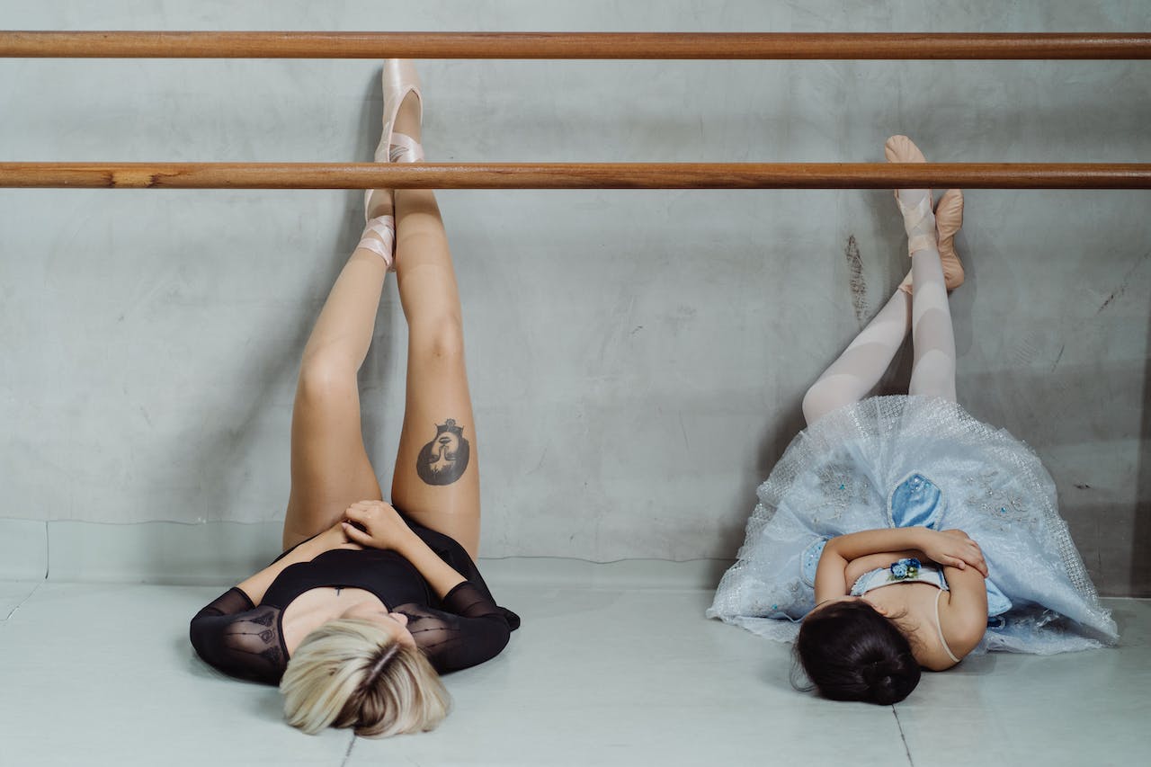 Dwie tancerki baletowe odpoczywają na podłodze, ich nogi wyciągnięte do góry i oparte o drążek do baletu, jedna w czarnym stroju z widocznym tatuażem na udo