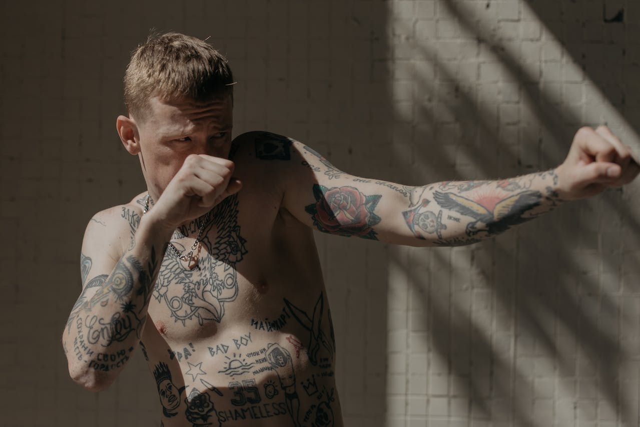 Mężczyzna w pozie bokserskiej eksponuje swoje liczne tatuaże na torsie i ramionach w jasno oświetlonym pomieszczeniu