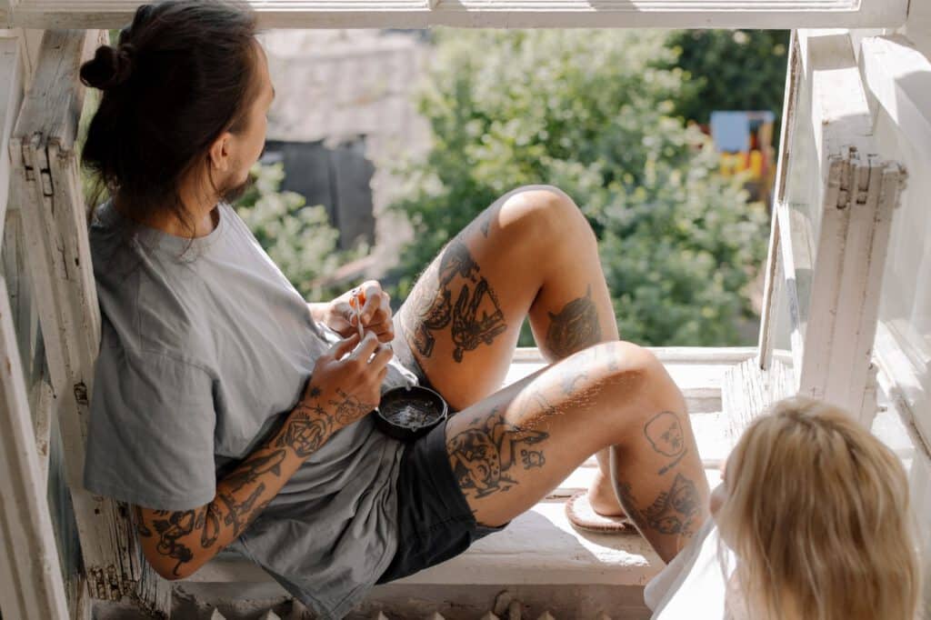 Siedzący na parapecie mężczyzna z tatuażami na nogach i rękach