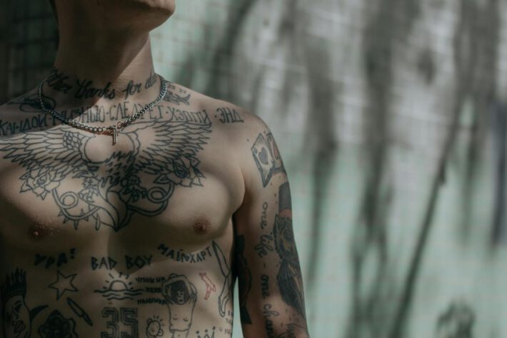 Tors mężczyzny pokryty jest wieloma tatuażami, w tym wyrazistym wizerunkiem rozpostartych skrzydeł na klatce piersiowej