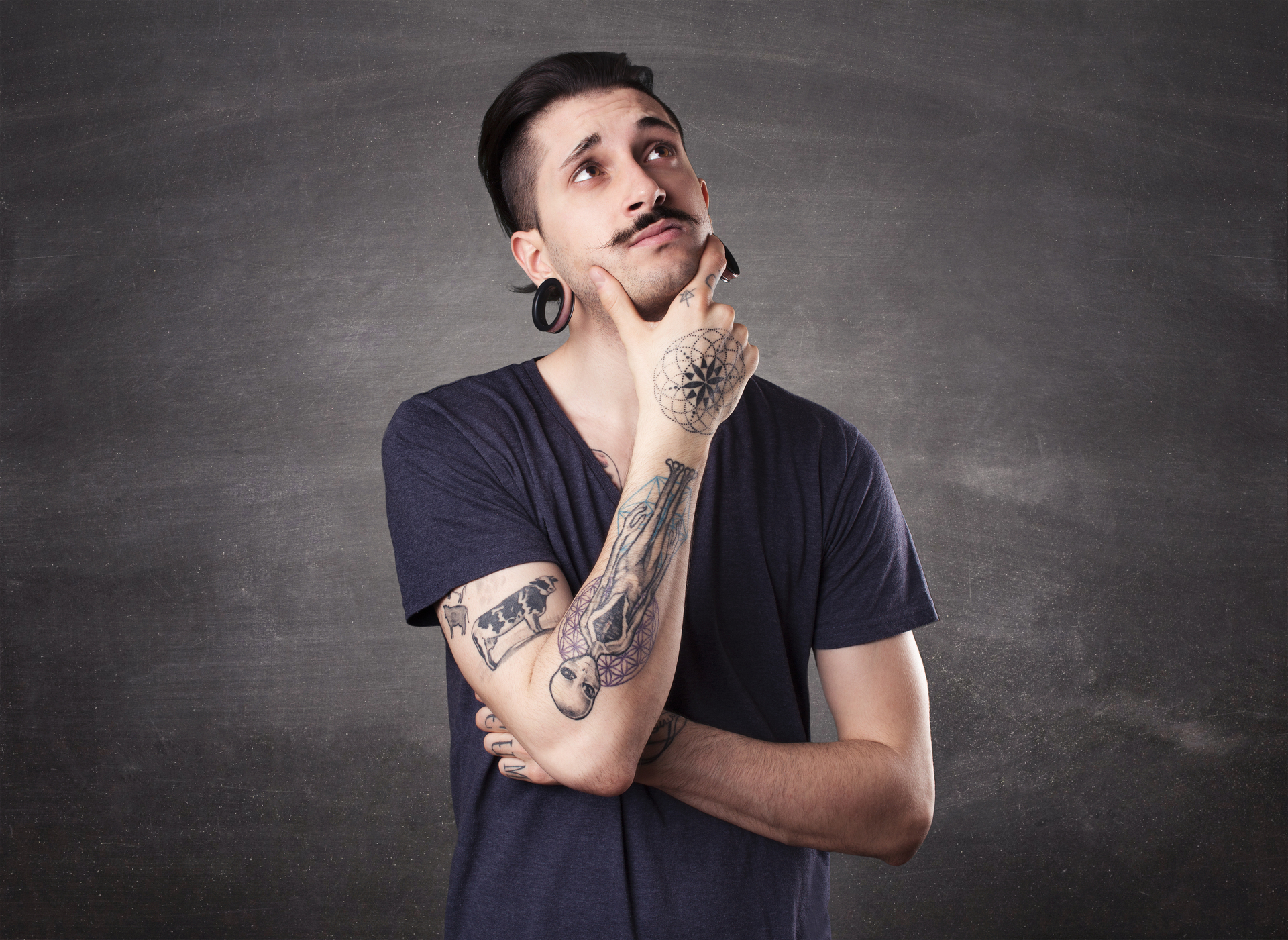 Mężczyzna z przekłutymi uszami i tatuażami na rękach zastanawia się, opierając podbródek o rękę z wyrysowanym na niej symbolem