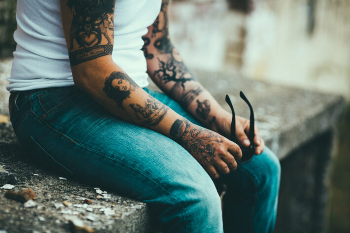 Osoba siedzi na kamiennym murku, trzymając w dłoniach okulary przeciwsłoneczne; jej ramiona zdobią liczne tatuaże o różnorodnych wzorach, w tym postaci i symbole