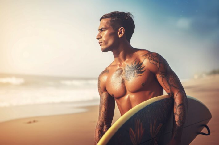 Mężczyzna o umięśnionej posturze stoi na plaży, trzymając deskę surfingową, z wyraźnym tatuażem o motywach roślinnych na klatce piersiowej i ramieniu