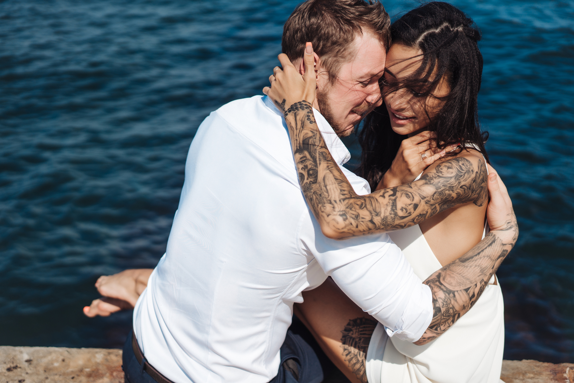 Para w bliskim objęciu wyraża czułość nad brzegiem morza, mężczyzna w białej koszuli i kobieta w białej sukni, oboje z tatuażami ozdabiającymi ich ramiona