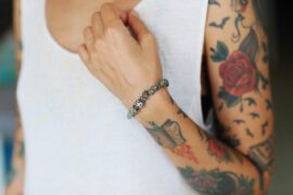 Kobieta z różnokolorowymi tatuażami na ramieniu nosi prostą bransoletkę z koralików. Na jej skórze widoczne są zarysy ptaków w locie, a także większy tatuaż przedstawiający czerwoną różę
