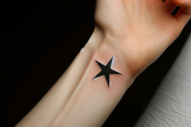 Na przedramieniu znajduje się tatuaż czarnej gwiazdy z delikatnym cieniowaniem, który wydaje się świeży, biorąc pod uwagę połysk na skórze. Tło jest jednolicie ciemne, co sprawia, że tatuaż jest centralnym punktem obrazu