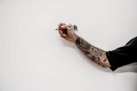 Wytatuowana ręka trzymająca maszynkę do robienia tatuażu, która dziarać będzie tatuaż na kostce