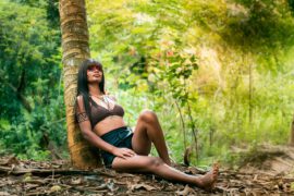 Kobieta o ciemnych włosach, ubrana w bikini, relaksuje się oparta o palmę wśród bujnej zieleni. Ma na ramieniu tatuaż w formie geometrycznych wzorów