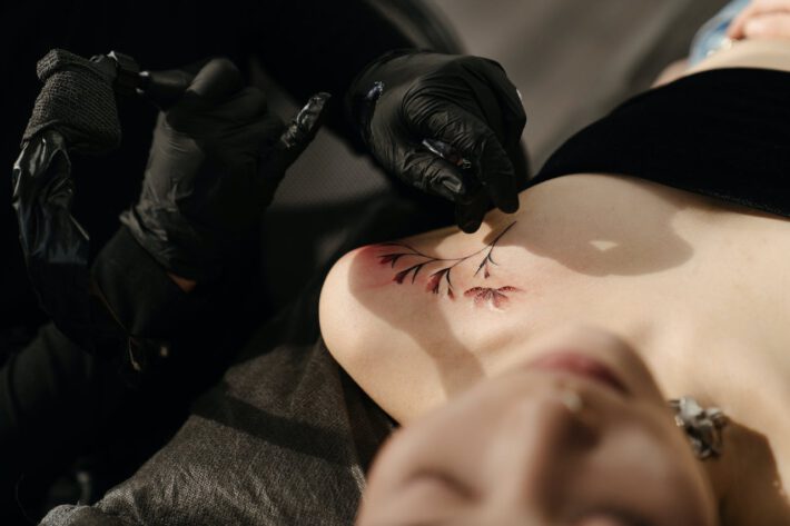 Osoba ubrana w czarne rękawiczki precyzyjnie wykonuje tatuaż w okolicy dekoltu leżącego klienta, przedstawiający kwiat z delikatnymi liniami