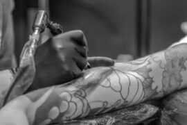 Artysta tatuażu pracuje nad dużym wzorem na ramieniu klienta. Widać jego rękawice i maszynkę do tatuowania, które tworzą kontrast z niedokończonym jeszcze tatuażem