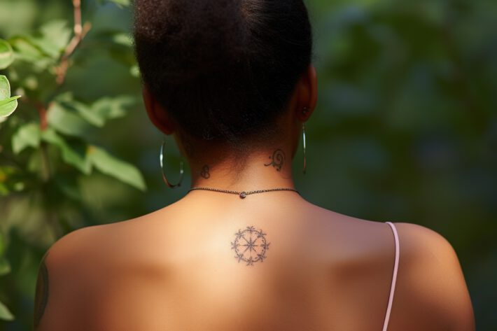 Kobieta stoi tyłem, eksponując tatuaż na karku przypominający kompas w otoczeniu dekoracyjnych motywów, a na jej skroniach widoczne są delikatne tatuaże