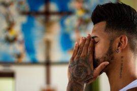 Mężczyzna z tatuażami na dłoni i szyi ma zamknięte oczy i złożone dłonie przy twarzy w geście modlitwy lub zadumy. Na jego szyi widnieje ciąg rzymskich cyfr