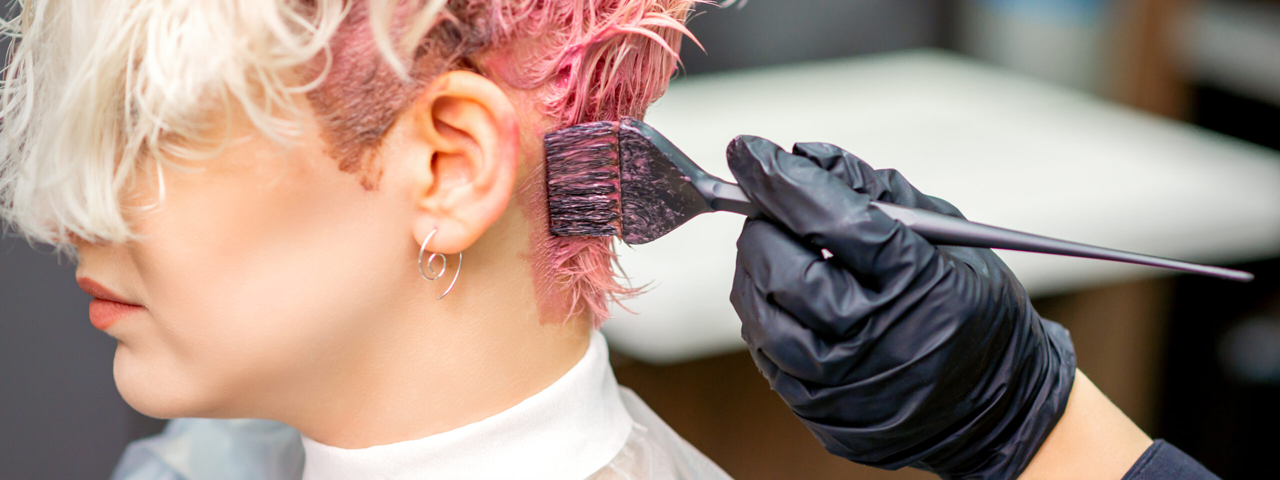 Na zdjęciu widać klientkę salonu fryzjerskiego, której fryzjerka farbuje krótkie włosy na różowo. 