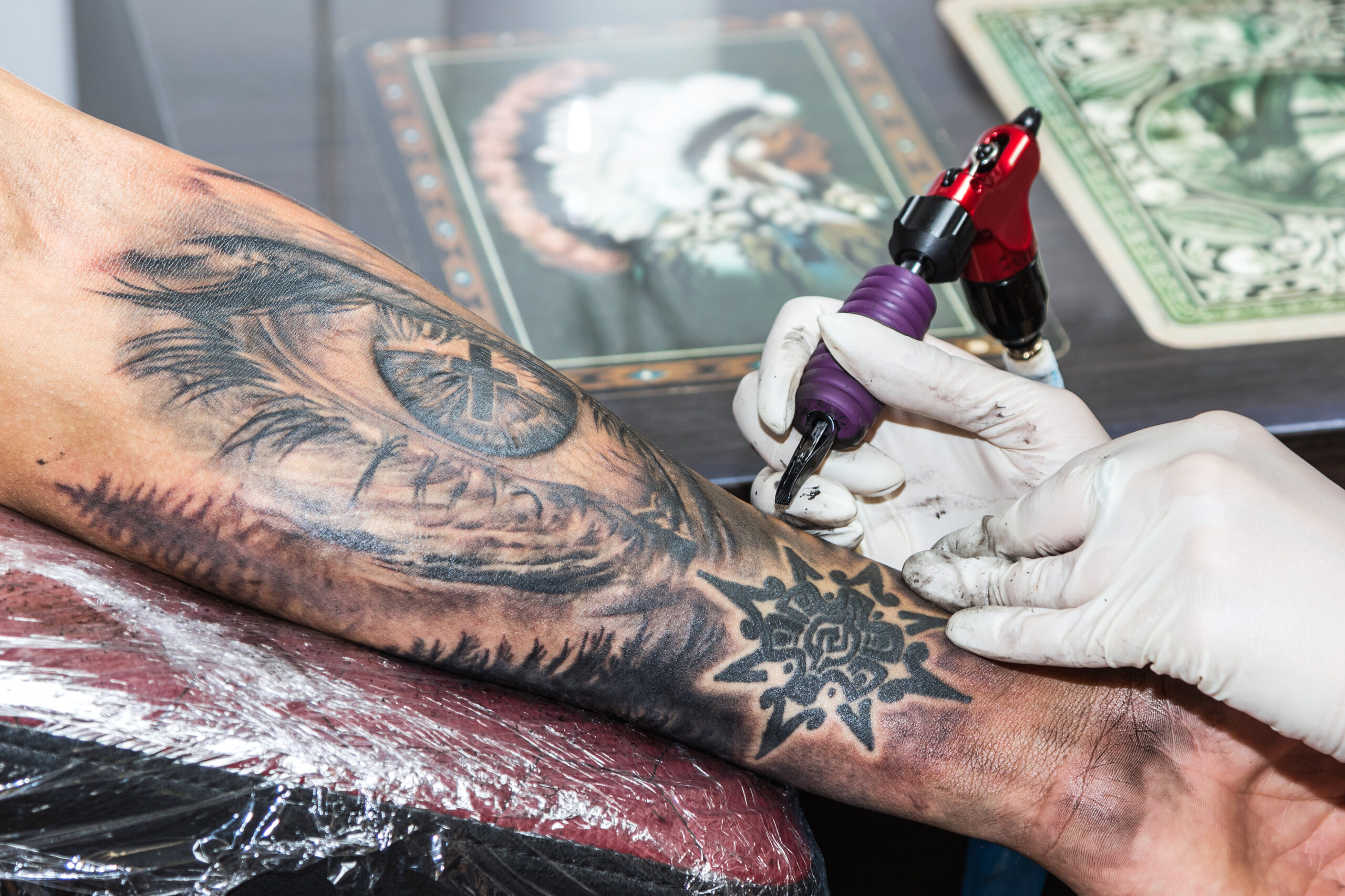 Na obrazku możemy zobaczyć artystę tatuażu, który tworzy imponujący męski tatuaż tribal na przedramieniu. Jego precyzyjne ruchy i umiejętności artystyczne są widoczne, gdy geometryczne wzory i linie zaczynają nabierać kształtu, nadając ramionom i nadgarstkom mocny i odważny charakter.