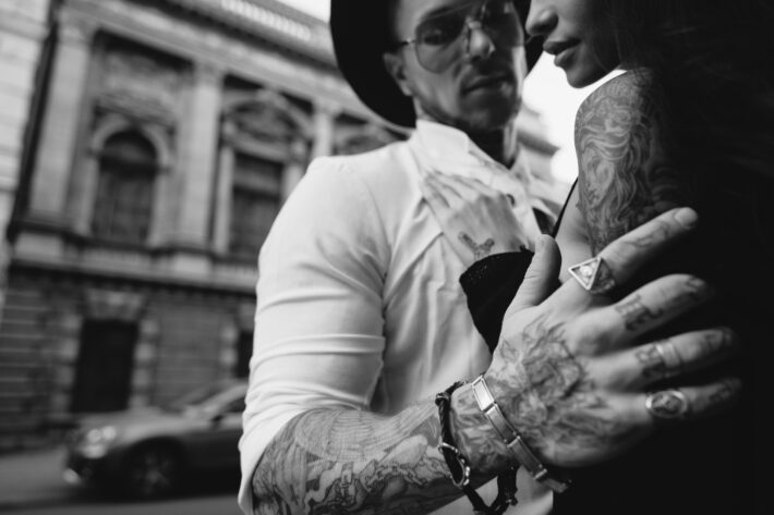 Na tym ujęciu widzimy mężczyznę trzymającego kobietę za ramiona na ulicy. Oboje mają tatuaże wykonane z okazji swoich czterdziestych urodzin. Obrazek utrzymany jest w czarno-białej tonacji, co podkreśla charakter i styl tatuaży. Mężczyzna i kobieta patrzą na siebie z uśmiechem, co podkreśla ich zadowolenie z wykonanych tatuaży oraz pewność siebie i swojego stylu. Obrazek ukazuje, że tatuaże to nie tylko ozdoba, ale także wyraz osobowości i indywidualnego stylu życia.