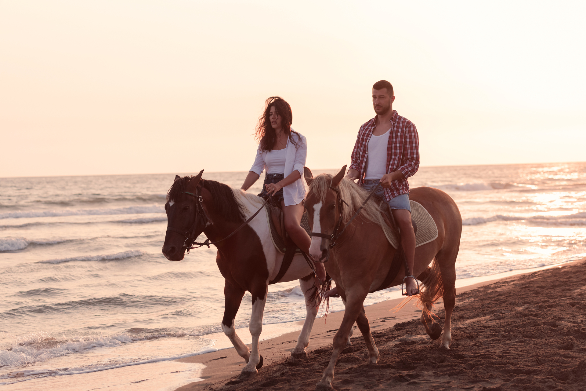 Na tym zdjęciu widzimy parę, która jeździ konno po plaży. Kobieta trzyma wodze swojego konia, a mężczyzna prowadzi swojego konia obok niej. W tle widać błękitne morze i piękny zachód słońca. Para wydaje się być bardzo zadowolona z przejażdżki i uśmiechnięta. Całość tworzy romantyczną i malowniczą atmosferę, która idealnie wpisuje się w letnie wakacje nad morzem.