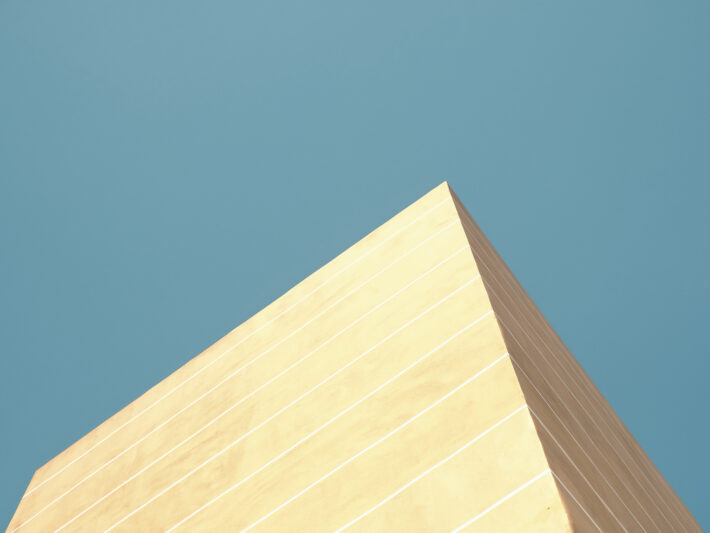 Ten malowniczy obrazek przedstawia część budynku o kształcie trapezu, wznoszącą się wśród pięknego tła nieba. Geometryczne linie trapezu harmonijnie kontrastują z niebem, tworząc efektowny widok. Obrazek emanuje nowoczesnością i solidnością, ukazując połączenie architektury i natury w niezwykłej kompozycji