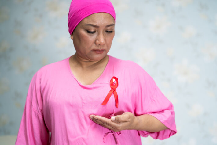 Na tym obrazku widzimy kobietę, która trzyma na głowie różową chustkę, symbolizującą walkę z rakiem piersi. Jej wyraz twarzy emanuje determinacją i siłą ducha w obliczu trudności. Przez tę symboliczną gestykulację wyraża swoje wsparcie dla wszystkich dotkniętych tą chorobą, dając nadzieję i inspirację innym
