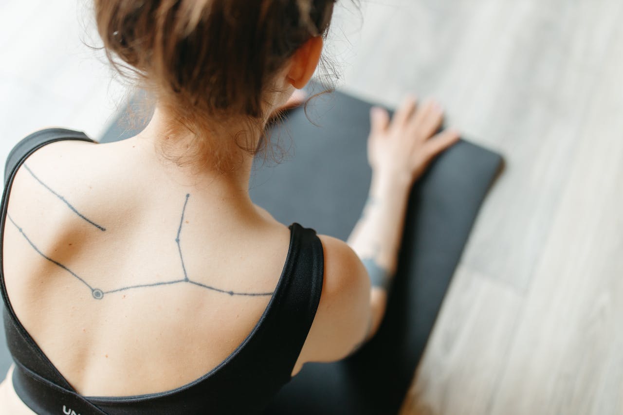 Kobieta z tatuażem przedstawiającym konstelację gwiazd na ramieniu przygotowuje się do ćwiczeń, opierając rękę na macie do jogi. Linie tatuażu są proste i czyste, tworząc minimalistyczny wzór na jej skórze. Ubrana jest w czarny, sportowy top, który odsłania jej ramiona i pozwala na pełny widok tatuażu. Włosy ma upięte, co dodatkowo eksponuje tatuaż na jej plecach