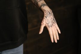 Tatuaż z misternym wzorem, który wygląda na azjatycką maskę demona lub mityczną postać, pokrywa rękę aż do palców. Ciemne linie tatuażu są wyraźne i kontrastują z jasną skórą, tworząc dramatyczny efekt