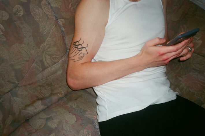 Osoba w białym bezrękawniku ma na przedramieniu tatuaż przedstawiający dwie dłonie, z których jedna delikatnie dotyka twarzy drugiej. Scena jest wykonana prostymi liniami, które tworzą subtelny i wyrafinowany obraz. Osoba ta trzyma w dłoniach telefon komórkowy, na którym coś czyta lub pisze, a jej postawa jest zrelaksowana, wygodnie oparta o tapicerowany mebel z wzorzystym obiciem. Na palcu widać obrączkę, co może sugerować, że jest to zarejestrowany związek lub małżeństwo