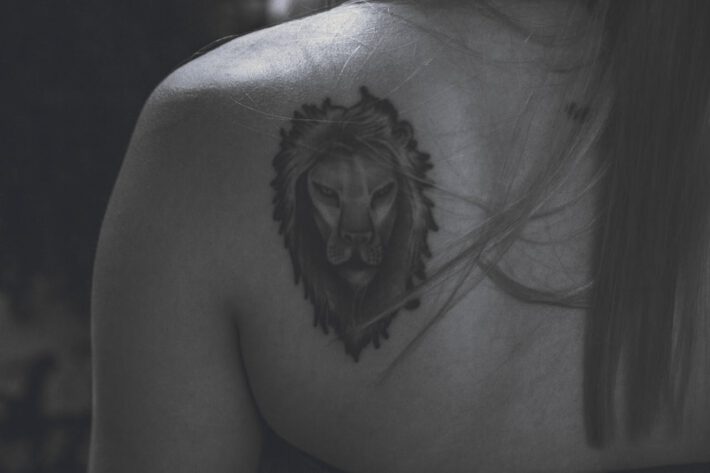 Na ramieniu pojawia się tatuaż głowy lwa, którego majestatyczna grzywa i przenikliwe spojrzenie nadają mu wygląd pełen godności i siły. Włosy spływające wzdłuż ramienia delikatnie przykrywają krawędzie tatuażu, co dodaje naturalności całej scenie