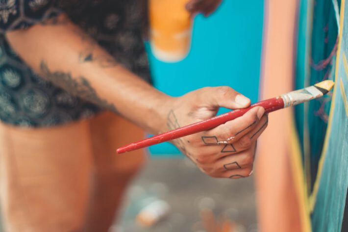 Ręka artysty, z widocznymi tatuażami na palcach i nadgarstku, trzyma czerwony pędzel przy malowaniu obrazu. Kontrast kolorów i tatuaż w geometryczne wzory przyciągają uwagę