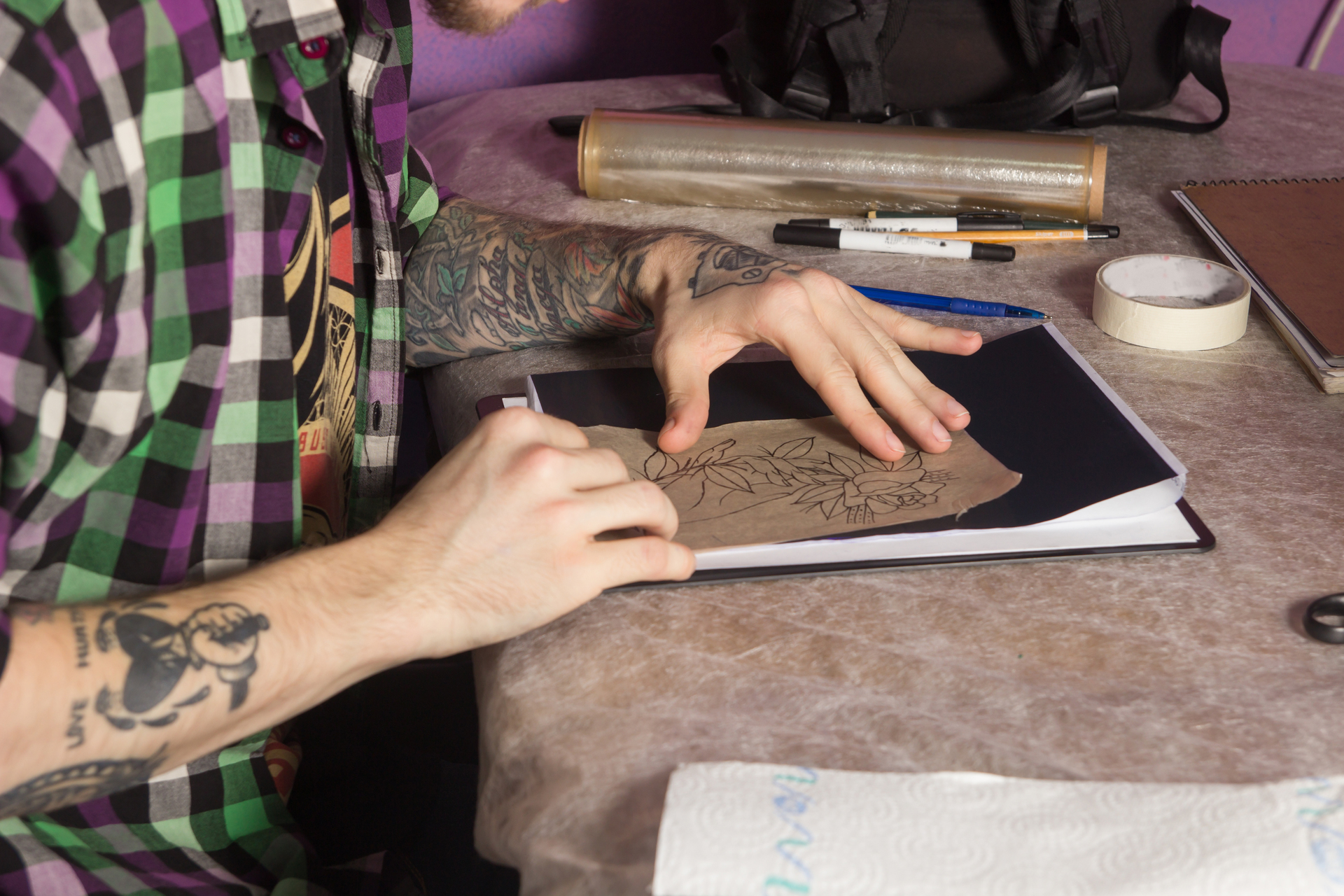 Obrazek przedstawia artystę tatuażu, który szkicuje swój nowy projekt na kartce papieru. Delikatne linie i precyzyjne kontury ożywiają koncepcję tatuażu, tworząc wstępne dzieło sztuki na płaskiej powierzchni. Ten moment twórczego procesu ilustruje umiejętności artysty, jego pomysłowość i zdolność do przenoszenia wizji na papier, przygotowując się do późniejszego etapu wykonania tatuażu na skórze