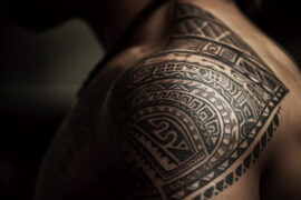 Szczegółowy tatuaż z motywami kultury rdzennych mieszkańców, pokrywający bark jak i plecy mężczyzny. Wzory są złożone i składają się z geometrycznych kształtów, spiralnych linii i tradycyjnych symboli, które są wyraźnie widoczne na ciemniejszym tle skóry. Oświetlenie akcentuje bogactwo detali tatuażu, nadając mu trójwymiarowy efekt