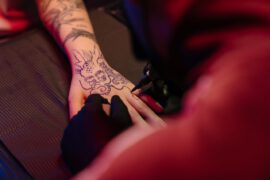 Zbliżenie na rękę osoby, która jest tatuowana przez tatuażystę. Projekt tatuażu, który wydaje się być skomplikowany i kwiatowy, jest konturowany niebieskim tuszem na skórze osoby. Widoczne są rękawiczki artysty, a otaczające światła rzucają ciepły, czerwony blask na scenę